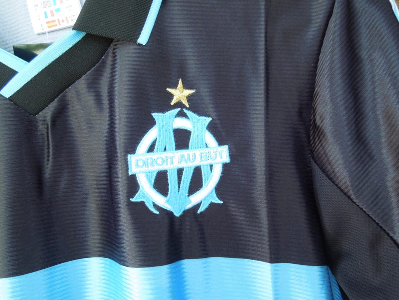 May include: A navy blue and light blue soccer jersey with the emblem of a team. The emblem features a light blue and white design with the words "DROIT AU BUT" and a gold star. The jersey has a V-neck collar.
