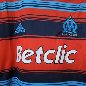 May include: Red and navy blue football shirt with light blue horizontal stripes. Includes the Adidas logo, the Betclic logo, and the Olympique de Marseille team emblem with the text "DROIT AU BUT".