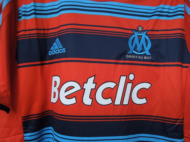 May include: Red and navy blue football shirt with light blue horizontal stripes. Includes the Adidas logo, the Betclic logo, and the Olympique de Marseille team emblem with the text "DROIT AU BUT".