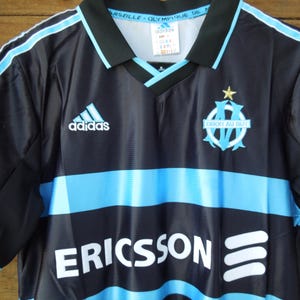 May include: Black and light blue soccer jersey with the Adidas logo and the Marseille team emblem. The jersey has a V-neck collar and the word "ERICSSON" printed across the front.