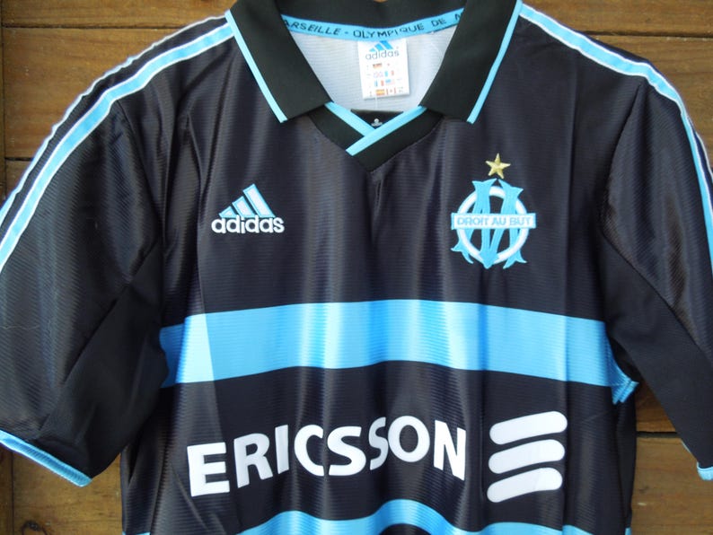 May include: Black and light blue soccer jersey with the Adidas logo and the Marseille team emblem. The jersey has a V-neck collar and the word "ERICSSON" printed across the front.