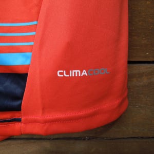 May include: Close-up of a red athletic shirt with blue horizontal stripes. The shirt has the text "CLIMACOOL" printed in silver on the sleeve. The fabric appears to be a breathable, athletic material.