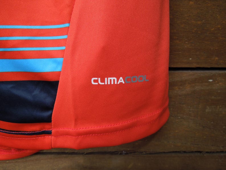 May include: Close-up of a red athletic shirt with blue horizontal stripes. The shirt has the text "CLIMACOOL" printed in silver on the sleeve. The fabric appears to be a breathable, athletic material.