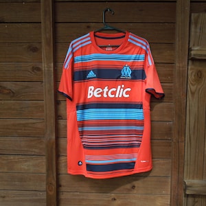 May include: Orange and blue striped football shirt with the Adidas logo and the text "Betclic". The shirt has a crew neck and short sleeves. The team logo is on the upper left chest.