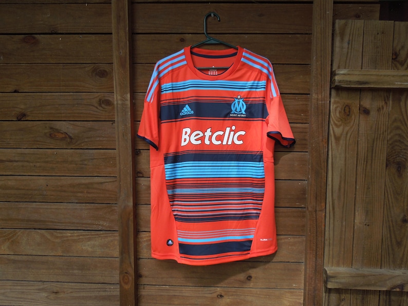 May include: Orange and blue striped football shirt with the Adidas logo and the text "Betclic". The shirt has a crew neck and short sleeves. The team logo is on the upper left chest.
