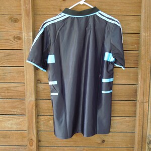 May include: Black athletic jersey with light blue accents. The jersey features light blue stripes on the shoulders and sides, and a black collar. The jersey is hanging on a white hanger against a wooden background.