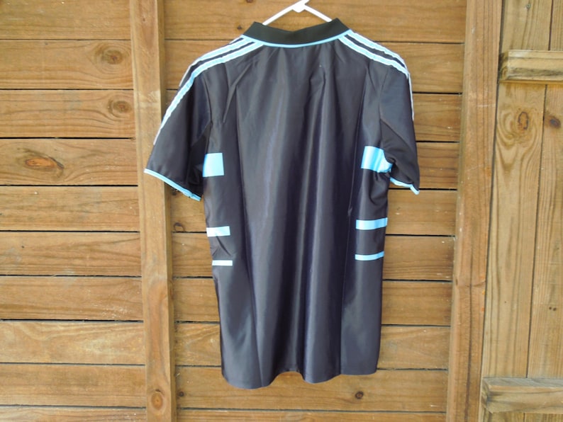 May include: Black athletic jersey with light blue accents. The jersey features light blue stripes on the shoulders and sides, and a black collar. The jersey is hanging on a white hanger against a wooden background.
