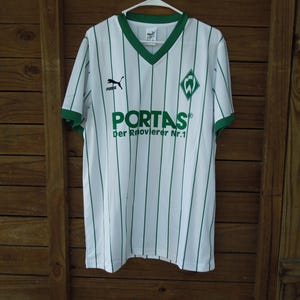 May include: A white and green striped football shirt with a V-neck collar. The shirt displays the Puma logo, a team emblem, and the word "PORTAS" in green. The text "Der Rekordmeister Nr.1" is also visible.