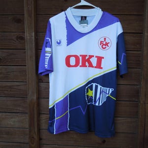 Könnte beinhalten: Ein weißes, lila und marineblaues Fußballtrikot mit dem Wort "OKI" in roten Blockbuchstaben. Das Trikot hat einen runden Kragen und kurze Ärmel. Es hat ein Logo auf der oberen linken Brust und eine Grafik unten rechts.