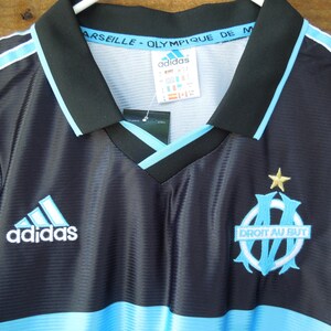 May include: Black and light blue soccer jersey with the Adidas logo and the Marseille team emblem. The jersey has a V-neck collar and light blue stripes on the shoulders. The text "Marseille - Olympique de M" is visible on the collar.