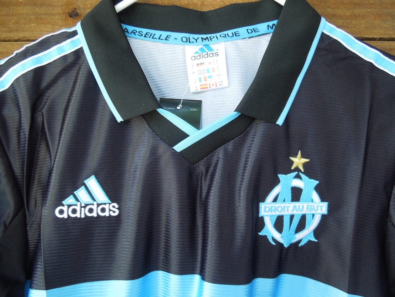 May include: Black and light blue soccer jersey with the Adidas logo and the Marseille team emblem. The jersey has a V-neck collar and light blue stripes on the shoulders. The text "Marseille - Olympique de M" is visible on the collar.