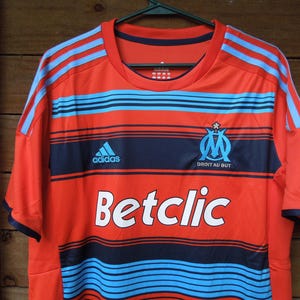 May include: Orange and navy blue football shirt with light blue stripes and the Adidas logo. The shirt features the text "Betclic" and the emblem of Olympique de Marseille.
