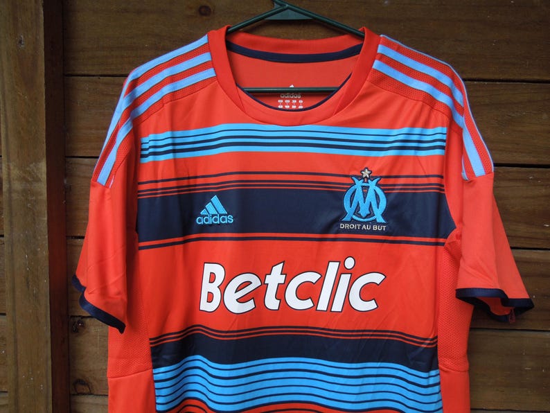 May include: Orange and navy blue football shirt with light blue stripes and the Adidas logo. The shirt features the text "Betclic" and the emblem of Olympique de Marseille.