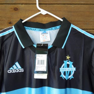 May include: Black and light blue Adidas soccer jersey with the Olympique de Marseille logo and text. The jersey has a black collar and light blue stripes on the shoulders. The Adidas logo is on the chest.