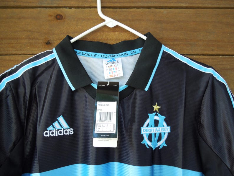 May include: Black and light blue Adidas soccer jersey with the Olympique de Marseille logo and text. The jersey has a black collar and light blue stripes on the shoulders. The Adidas logo is on the chest.