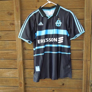 May include: Black and blue Adidas soccer jersey with the word "ERISSON" across the front. The jersey has light blue stripes and accents, a collar, and the Adidas logo. The team logo is on the chest.