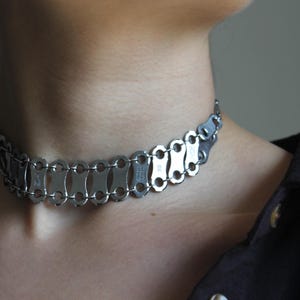 Puede incluir: Un collar gargantilla plateado y un juego de pendientes a juego. El collar y los pendientes están hechos de piezas de metal entrelazadas, creando un diseño industrial único. La joya se lleva con una camisa oscura.