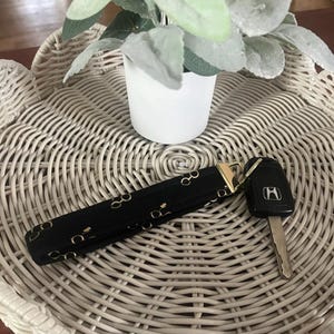 Puede incluir: Llavero negro con estampado de gafas doradas y cierre dorado. Se adjunta una llave de coche negra con una hoja plateada. El llavero está sobre una bandeja de mimbre blanca con una maceta blanca pequeña y una planta verde.