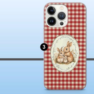 May include: A phone case with a red and beige gingham pattern. The case features an oval illustration of three bunnies. The bunnies are light brown and beige, with a white border around the illustration.