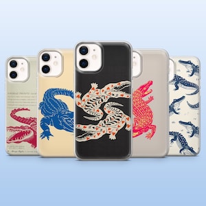 Op de afbeelding: Een verzameling telefoonhoesjes met alligatorontwerpen. De hoesjes tonen verschillende illustraties, waaronder een rode alligator, een blauwe alligator en een zwarte hoes met witte en rode alligatorkunst. De hoesjes zitten in doorzichtige beschermhoezen.