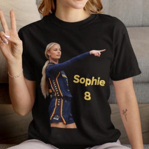 Può includere: Una t-shirt nera a maniche corte con la grafica di un'atleta bionda in divisa da basket blu navy e gialla che indica verso destra. Il testo "Sophie" e il numero "8" sono stampati in giallo sul lato destro della grafica.