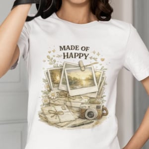 Pode incluir: Uma t-shirt branca de manga curta com uma impressão gráfica de fotos polaroid empilhadas, uma câmara vintage e uma carta com um laço. O texto "MADE OF HAPPY" está impresso em castanho escuro acima das ilustrações, rodeado por detalhes florais.