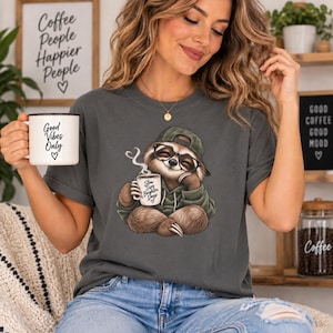 May include: A charcoal grey t-shirt featuring a graphic of a sloth wearing a green hoodie, a baseball cap, and glasses, holding a mug with the text "Slow Sips Brighter Days". The sloth is sitting in a relaxed pose with steam rising from the mug.
