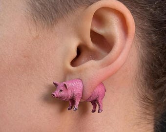 Puede incluir: Un pendiente con forma de cerdo rosa que parece atravesar el lóbulo de la oreja, con la parte delantera del cerdo en la parte frontal y la parte trasera en la parte posterior, creando el efecto de una figura de cerdo continua.