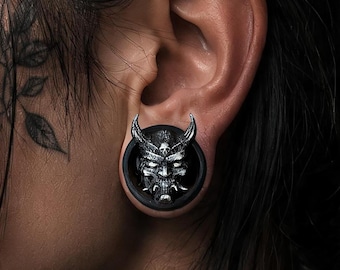 Puede incluir: Un expansor de oreja circular con un diseño detallado de máscara de demonio u oni en tono plateado, con cuernos prominentes y dientes afilados, sobre un fondo negro sólido. La pieza de joyería se lleva en el lóbulo de la oreja.
