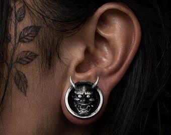 Puede incluir: Un expansor de oreja circular con un diseño detallado de máscara Hannya en tono plateado, con cuernos prominentes, una sonrisa amplia y dientes afilados. El accesorio metálico tiene un acabado oscuro y desgastado en un marco pulido.