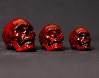 Puede incluir: Tres figuras de cráneos de color rojo intenso brillante dispuestas en fila de mayor a menor. Cada cráneo presenta cuencas oculares detalladas, cavidad nasal y una mandíbula definida con dientes visibles, todo en un acabado carmesí sólido.