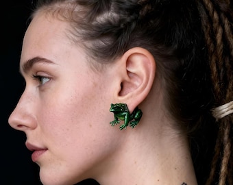 Puede incluir: Un pequeño dilatador o pendiente de oreja con forma de rana de color verde oscuro que se lleva en el lóbulo. La figura de la rana está detallada con extremidades visibles y una superficie texturizada, como un adorno tridimensional en la oreja.