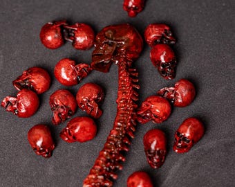 Puede incluir: Una colección de pequeñas figuras de cráneos de color rojo oscuro brillante esparcidas alrededor de una pieza central de columna vertebral vertical. El conjunto consta de quince cráneos individuales y una columna vertebral segmentada, todos con un acabado carmesí.