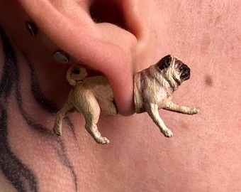 Puede incluir: Una pieza de joyería para oreja con forma de perro pug en color beige y negro, diseñada para parecer que un perro atraviesa el lóbulo. La sección frontal muestra la cabeza y las patas delanteras, mientras que la trasera muestra las patas traseras.