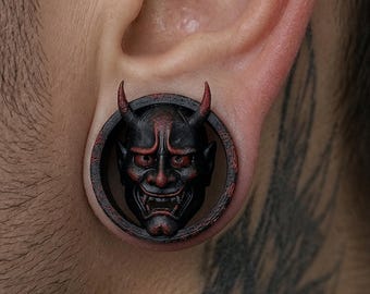 Puede incluir: Un dilatador de oreja circular que presenta una máscara de demonio negra y roja detallada con cuernos prominentes. La máscara está centrada dentro de un marco de anillo oscuro y desgastado, mostrando rasgos faciales intrincados, dientes afilados y acentos de color rojo oscuro en los ojos y los cuernos.