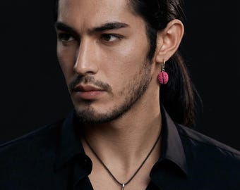Puede incluir: Un pendiente de aro de plata y un collar con colgante a juego, ambos con un pequeño dije texturizado de color rosa brillante con forma de cerebro. El conjunto de joyas se lleva con una camisa negra lisa, resaltando el color vibrante y el diseño.