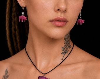 Puede incluir: Un conjunto de joyería compuesto por dos pendientes colgantes y un collar con colgante. Cada pieza presenta una pequeña figura de cerdo de color rosa brillante. Los pendientes cuelgan de ganchos plateados y el colgante está en un cordón negro.