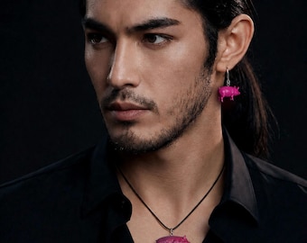 Puede incluir: Un conjunto de joyería que consta de un pendiente individual y un collar, cada uno con una pequeña figura de cerdo de color rosa brillante. El pendiente es de estilo colgante con un gancho plateado y el collar tiene un cordón negro.