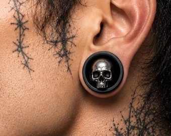 Puede incluir: Un expansor de oreja circular con un borde exterior negro y un diseño de calavera en tono plateado en el centro. La calavera presenta cuencas oculares huecas y una cavidad nasal definida, sobre un fondo oscuro dentro del marco circular.