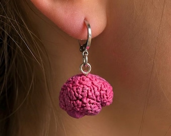 Puede incluir: Un pendiente de aro plateado con un pequeño dije texturizado de color rosa brillante con forma de cerebro humano. El dije cuelga de una anilla metálica circular que atraviesa el lóbulo de la oreja, mostrando una textura superficial detallada.