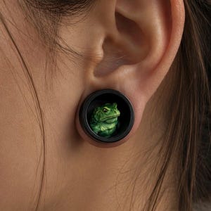 May include: A circular ear plug featuring a black rim and a detailed, three-dimensional green toad figurine centered within the opening. The toad has textured skin, visible eyes, and a realistic posture, set against a dark interior background.