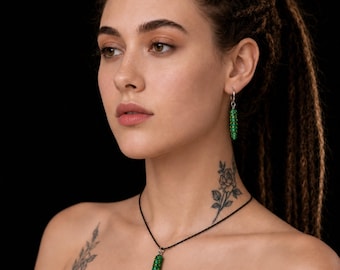 Puede incluir: Un conjunto de joyería que consta de un collar con colgante y un pendiente individual. El colgante es un pequeño objeto cilíndrico verde con textura que cuelga de un cordón negro fino. El pendiente presenta un dije cilíndrico verde a juego.