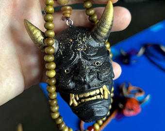 Puede incluir: Un colgante de máscara Hannya negro con cuernos y dientes pintados en oro, suspendido de un collar de pequeñas cuentas de madera redondas de color verde oliva. La máscara presenta una superficie texturizada con sutiles motas doradas.