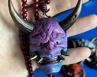 Puede incluir: Un colgante que presenta una máscara oni de color morado y azul con grandes cuernos texturizados de color bronce y colmillos afilados. La máscara está unida a un collar largo hecho de pequeñas cuentas redondas de color rojo oscuro.
