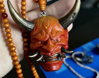 Puede incluir: Un colgante que presenta una máscara de demonio roja y naranja con cuernos grandes, oscuros y texturizados y colmillos metálicos, suspendido de un largo collar de cuentas de madera de color marrón claro, lisas y redondas.