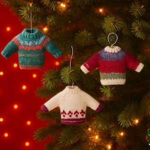 May include: Three miniature knitted sweater ornaments in teal, red, and white hang from a Christmas tree. The sweaters have colorful patterns and are suspended by silver hooks. The background is a festive red with soft, glowing lights.