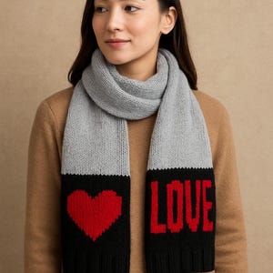 May include: A gray knit scarf with black ends featuring a red heart and the word "LOVE" in red block letters. The scarf is draped around a person's neck, with a brown background.