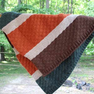 Puede incluir: Una manta de punto con rayas diagonales en tonos naranja, marrón, beige y verde. La manta está colocada sobre una barandilla de madera, con un bosque visible al fondo. La textura de la manta es visible.