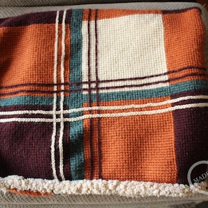 May include: A woven blanket featuring a plaid pattern in shades of burnt orange, deep burgundy, teal, and cream. The blanket has a textured waffle-like weave and is finished with a cream-coloured fringe border. Text in the corner reads MADE WITH LOVE AND BAD WORDS Corri.