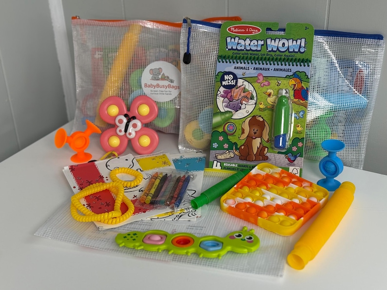 May include: A collection of colorful children's toys, including a butterfly-shaped toy, a water-reveal activity book, and a pop-it toy. The toys are arranged on a white surface, with some items inside clear mesh bags. The color palette includes orange, pink, yellow, and green.
